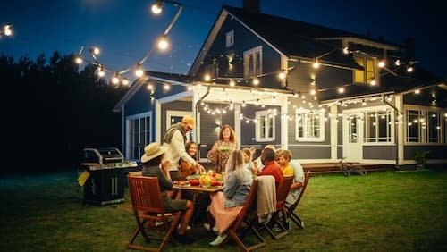 family and friends sitting around outdoor table with food next to house in evening