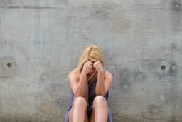 a young woman with her back against the wall sitting on the ground with her head in her hands