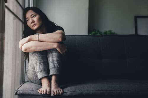 woman sitting on sofa with knees pulled into her chest, depressed and sad, looking out the window